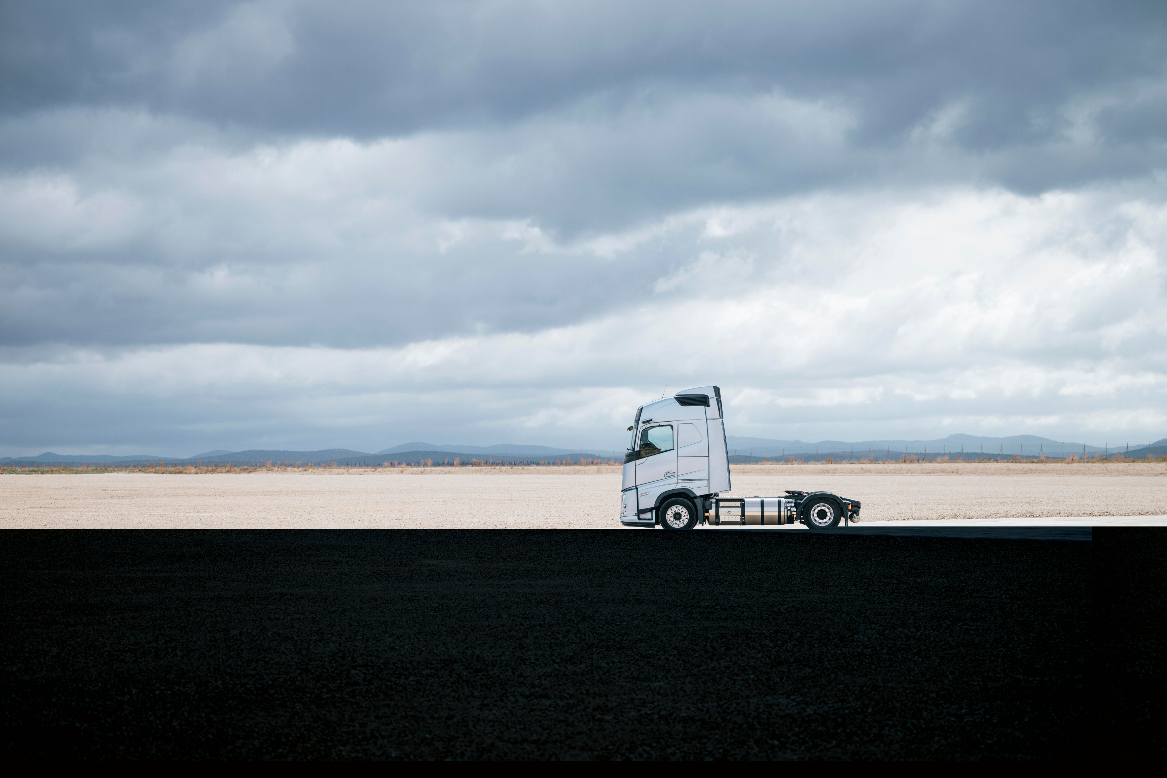 Страничен изглед на камион Volvo, паркиран на широк, открит терен под облачно небе, с обширна околност и минималистичен пейзаж.